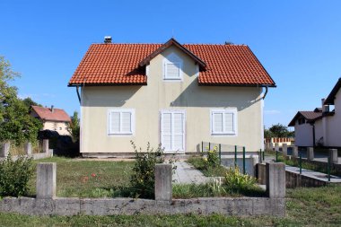Krem rengi çatı, beyaz panjurlar ve küçük yatakhane beton direklerle çevrili çimenli bir bahçede ve açık mavi gökyüzünün altında komşu evlerle çevrili.