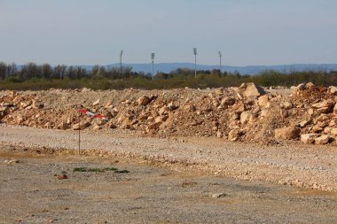 Bir inşaat sahasında büyük bir kayalık toprak yığını, ön planda kırmızı ve beyaz çizgili bir işaret, uzak ağaçlar ve stadyum ışıkları berrak bir gökyüzünün altında görünür.