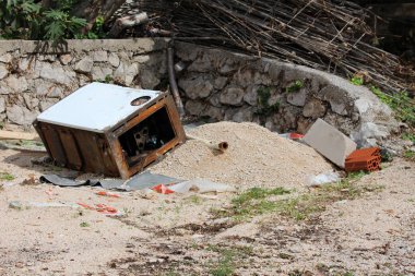 Paslı metal eşyalar, taş duvarlar, kum yığınları ve dağınık bina atığıyla çevrili bir inşaat sahasında, çakıl, tuğla ve molozların arasında yan yatar.