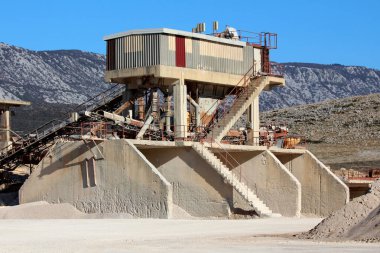 Yüksek kontrol odası, çoklu taşıma bantları ve uzun merdivenleri olan büyük endüstriyel beton işleme tesisi, açık mavi gökyüzünün altındaki uzak dağlarla kayalık bir arazide kurulmuş.