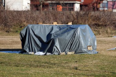 Gri bir muşambanın altına gizlenmiş, kenarları taşlarla kaplı büyük bir nesne, arka planda kuru bitki örtüsü ve binaları bulanık, yamalı bir çim tarlasında duruyor.