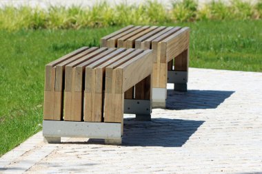 Paralel kirişli ve metal destekli iki minimalist ahşap bank, halka açık bir parkta yeşil çimenler ve süs bitkileriyle çevrili, güneşli taş kaldırımlı bir yolda konumlandırılmıştır.