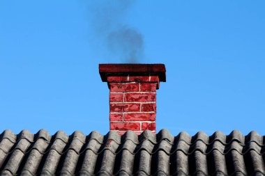 Kırmızı tuğlalı baca, açık mavi gökyüzüne keskin bir şekilde çizilmiş kıvrımlı beton çatıdan siyah duman yayar. Bu da yerleşim bölgesinde aktif ısıtma veya yanma olduğunu gösterir.