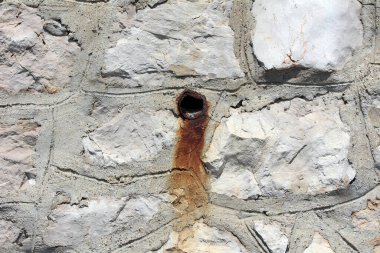 Üzerinde küçük paslı metal bir boru bulunan eskimiş taş duvar, altında turuncu-kahverengi bir pas lekesi bırakıyor, sert açık renkli taşlar ve gri havan topu ile çevrili.