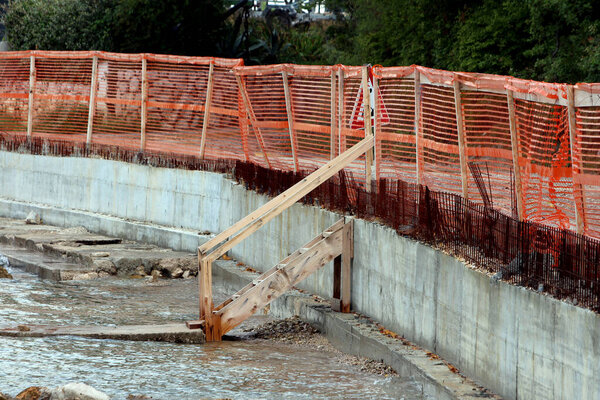 Защищающая бетонная стена с оранжевым пластиковым ограждением проходит вдоль береговой линии, с открытой деревянной подпоркой и арматурой, отделяющей строительную площадку от воды