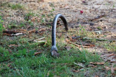 Topraktan çıkan kavisli bir metal boru, çimenlerin ve dağınık ahşap döküntülerinin arasından muhtemelen eski bir kamu parkının demirleme noktasını işaret ediyor.