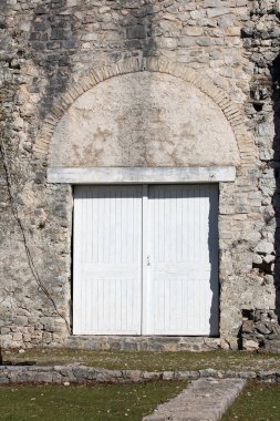 Tarihsel taş duvar kapalı çift beyaz ahşap kapıların üzerinde kemerli bir lintel içeriyor, kırsal duvarları klasik mimari detaylarla harmanlıyor.