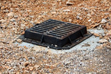 Sağlam bir siyah metal kanalizasyon ızgarası çakıllarla çevrili beton bir çerçevenin içinde duruyor. Yerdeki drenaj girişini emniyete alıyor.