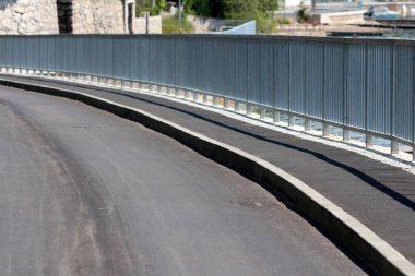 Pürüzsüz siyah asfalt yol, taştan duvarlar ve açık güneşli gökyüzü ile rıhtımda gezinen demir güvenlik korkuluklarının yanında yavaşça kıvrılır.
