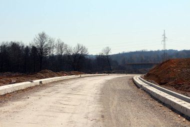 Ağaçlık ve uzak tepelerle çevrili bir inşaat alanından geçen yeni yapılmış beton kaldırımlarla süpürülen bir çakıl yatağı.