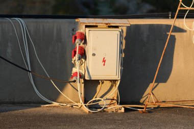 Kırmızı endüstriyel prizleri ve birbirine dolanmış kabloları olan yıpranmış bir açık hava elektrik dolabı beton bir duvarda duruyor, sıcak akşam güneşiyle yıkanmış.