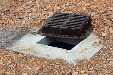 Kara metal bir rögar ızgarası çakıl kaplı bir zeminde hafifçe aralıktır. Beton bir çerçevenin altında karanlık bir drenaj açılır.