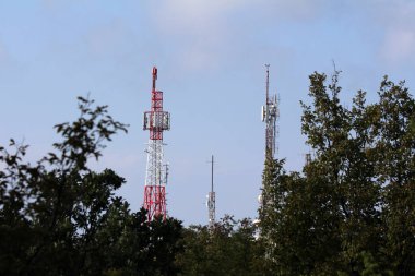 Antenlerle donatılmış çoklu kırmızı ve beyaz metal iletişim kuleleri ve yoğun yeşil ağaç tepelerinin üzerinde yükselen kaplar, açık mavi gökyüzüne karşı kurulmuş, modern kablosuz altyapıyı gösteriyor