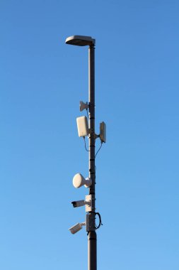 Uzun bir sokak lambası direği güvenlik kameraları, antenler ve kablosuz vericilerle donatılmıştır. Bulutsuz mavi bir gökyüzüne karşı durarak kentsel gözetim teknolojisini ön plana çıkarırlar.