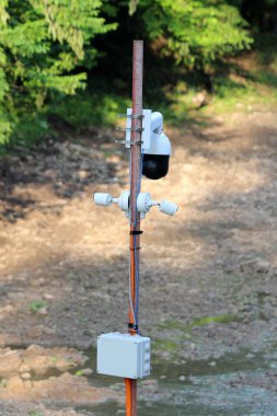 Paslı bir direğe, arka planda sık ağaçlar bulunan bir ormanı izleyen çok sayıda güvenlik kamerası ve elektrik kutusu yerleştiriliyor.