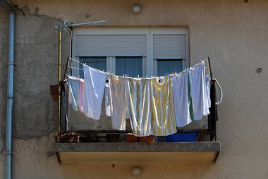 Çeşitli renk ve desenlerde çamaşırhaneler balkon korkuluklarından sarkıyor, pencerenin altındaki yıpranmış beton ve saksı bitkilerinin üzerine gölgeler döküyor.