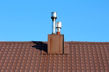 İki modern metal baca kahverengi kiremitli bir çatıdan yükseliyor, bulutsuz mavi gökyüzüne keskin bir şekilde çizilmiş, kullanım ve mimari tarzın bir karışımını gösteriyor