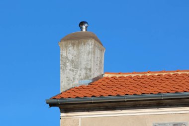 Modern metal kapaklı, çağdaş beton baca klasik terracotta çatı kiremitlerinin üzerine yükselir, açık mavi gökyüzü altında mimari dönemlere zıt düşer.