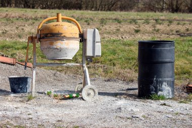 Kullanılmış sarı bir beton karıştırıcı, arka planda yeşil çimenler ve dağınık şişeler bulunan çakıl üzerinde siyah bir fıçı ve kova yanında duruyor.