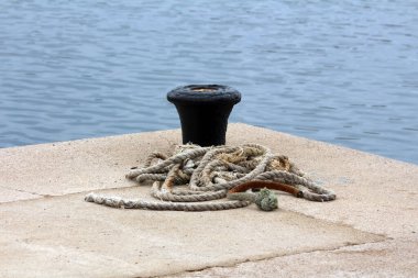 Yıpranmış bir halat, güneşli beton bir iskelede siyah bir kordonun yanında uzanır. Durgun mavi sulara bakar, deniz sanayisini ve sakin liman hayatını anımsatır.