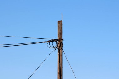 Yıpranmış bir ahşap elektrik direği, kırsal altyapı ve enerji dağılımını ön plana çıkaran parlak mavi gökyüzüne bağlı karmaşık elektrik kablolarından oluşan bir ağı destekler.