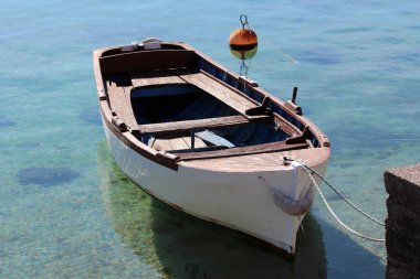 Kırsal ahşap bir kayık, boyası soyulmuş şeffaf turkuaz suda yüzer, beton bir iskelenin yanında görünür turuncu bir şamandıra ile bağlanır.