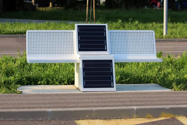 Entegre güneş panellerine sahip modern beyaz bank, halka açık alanlarda sürdürülebilir oturma ve cihaz şarj sunan yemyeşil çimlerin yanında kaldırımda duruyor.