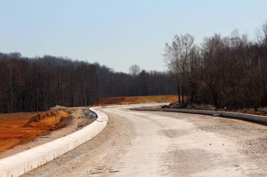 Yeni inşa edilmiş, beton bariyerlerle dolu kıvrımlı bir yol. Ağaçlar ve kırmızı toprakla çevrili, parlak, bulutsuz bir gökyüzünün altında ormanlık bir alanda esiyor.