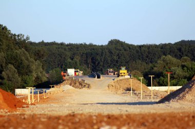 Kısmen inşa edilmiş bir otoyol, açık bir gökyüzünün altında çakıl ve toprak setleri üzerinde çalışan buldozerler, silindirler ve kamyonlarla bir orman boyunca uzanır.