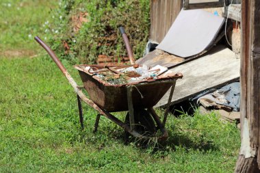 Yıpranmış, paslı bir el arabası inşaat atıkları ve bitki küpürleriyle dolup taşmış. Parlak güneş ışığı altında harap olmuş bir ahşap barakanın yakınındaki çimlerin üzerinde duruyor.