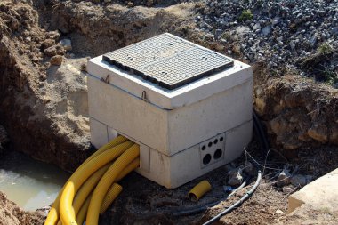 Bir inşaat sahasında kısmen gömülü olan beton bir elektrik bağlantı kutusu yan tarafına giren çoklu sarılı kanal boruları aktif yeraltı kablo kurulumunu gösteriyor.