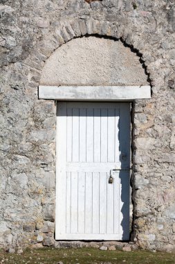 Metal bir asma kilitle korunan sağlam beyaz ahşap bir kapı, parlak güneş ışığıyla aydınlatılan kemerli bir kirişle sert bir taş duvara konur.