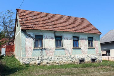 Şiddetli duvar çatlakları, mavi-yeşil boya soyulan küçük bir kırsal ev ve sarkık kiremitli çatı terk edilmiş bir köyde yapısal bozulmayı ve terk edilmeyi yansıtıyor.