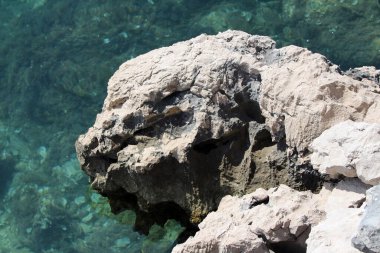 Engebeli bir kireçtaşı, şeffaf mavi suyun üzerine çıkar, yıpranmış yüzeyi keskin açılar ve doğal yarıklar gösterir, kıyı jeolojisinin saf güzelliğini anımsatır.