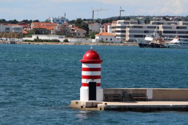 Küçük kırmızı ve beyaz çizgili deniz feneri, arka planda binalar, tekneler ve kırmızı çatılardan oluşan mavi sulara tepeden bakan beton bir iskelenin sonunda duruyor.