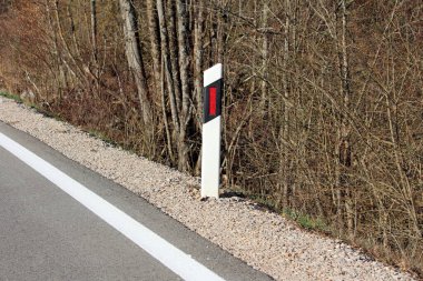 Kırmızı işaretli beyaz bir yol kenarı delinatör direği, yapraksız ağaçlar ve kuru çalılarla çevrili kırsal bir yolun yanındaki çakıl taşlı bir omuz üzerinde dikilir.