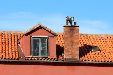Çatlak camları olan yıpranmış bir yatakhane penceresi ve güneşli terasotta kiremitli çatıdan yükselen tuğladan baca canlı renkte Avrupa konut mimarisini resmediyor.