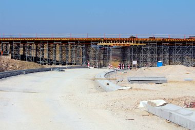 Geniş bir inşaat iskelesi kullanılarak büyük bir köprü inşa ediliyor. Toprak işleri, yol hazırlıkları ve açık mavi gökyüzü altında görünen inşaat malzemeleri büyük bir sivil proje üzerinde.