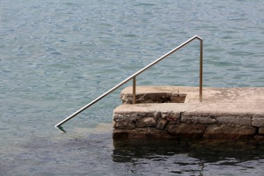 Sade bir taş ve beton iskele sakin bir günde yüzmek ya da kayıkla gezmek için sessiz bir giriş noktasını sembolize eden paslanmaz çelik bir tırabzanla donatılmış durgun bir suya dönüşür.