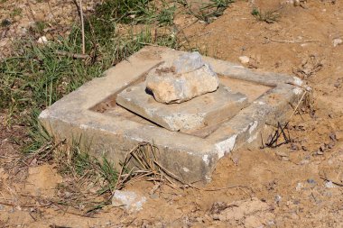 Toprak ve çimlerle çevrili boş bir kare beton temeli, tepesine taşlar yerleştirilmiş, yakın zamanda kaldırılmış bir yol yapısı veya elektrik direğinin kanıtlarını gösteriyor.
