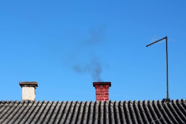 Kırmızı tuğlalı baca gri kıvrımlı bir çatıdan siyah dumanı gökyüzüne gönderirken, diğer bir baca ve televizyon anteni de yakınlarda duruyor.