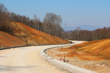 Beton bariyerlerle döşenmiş, kıvrımlı bir gelecek yolu taze kesilmiş ve turuncu toprağı aşıyor, ormanların kıyısındaki toprak ve toprak setler arasında kıvrılıyor.