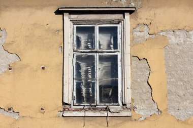 Beyaz boyanın soyulduğu eski bir ahşap pencere, yıkık dökük sarı alçı bir duvarın içinde duruyor. Bu da binaların onlarca yıllık yıpranma ve ihmalkârlığını ortaya çıkarıyor..