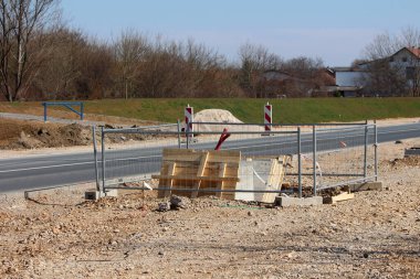 Yol kenarındaki bir inşaat alanında derin bir kazı, ahşap işleme ve yol güvenliği için gerekli borularla çevrili geçici metal bir çit yer alıyor.