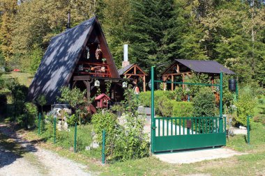 Koyu renk ahşap kaplamalı güzel bir kulübe, iki ahşap çardak ve modern yeşil bir kapı eşliğinde yemyeşil bir bahçeye kurulmuş.