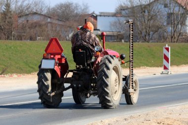 Kamuflaj ceketi ve turuncu başlıklı bir çiftçi, yeşillikle çevrili, güneşli kırsal asfalt bir yolda, çim biçme makinesi ile donatılmış klasik bir kırmızı traktörü kullanıyor.