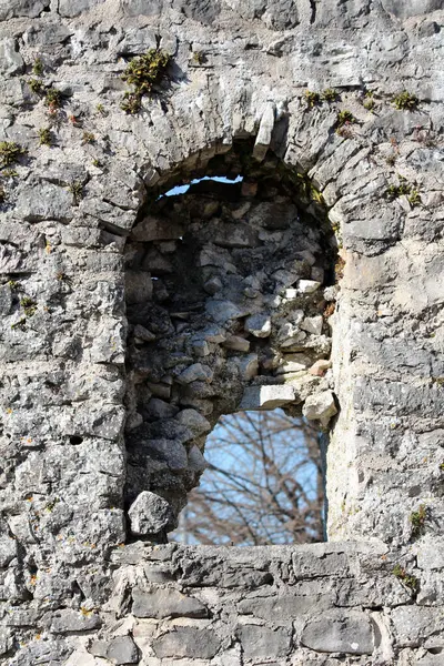 Eski bir duvarda, yıkılmış bir taş pencerenin yakın çekimi. Yıkılan duvarlar ve seyrek yosunlar. Çerçeveler çıplak ağaç dalları ve gökyüzü, tarihsel bozulmayı çağrıştırıyor.