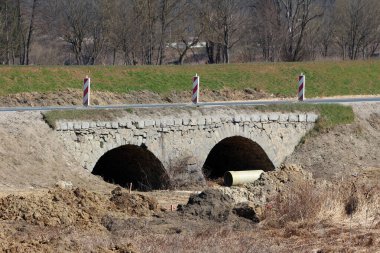 Tarihi çift kemerli taş köprü, bitişik yol boyunca görünen yol çalışması ve güvenlik işaretleriyle kurumuş, kazılmış bir nehir yatağını kaplıyor.