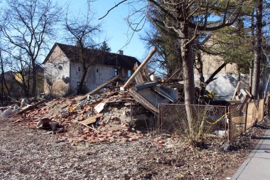 Bir yığın tuğla, ahşap ve kırık camlar yıkılmış bir binanın kalıntılarını işaret ediyor, yakınlardaki evler ve ağaçlar el değmeden durdukça enkaz toprak boyunca yayılıyor.