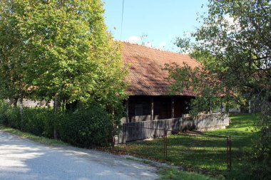 Kırsal ahşap, kırmızı kiremit çatılı bir ev ağaçlarla gölgelenmiş ve çitlerle çevrili. Geleneksel kırsal mimariyle yeşil yaz büyümesini harmanlamış.